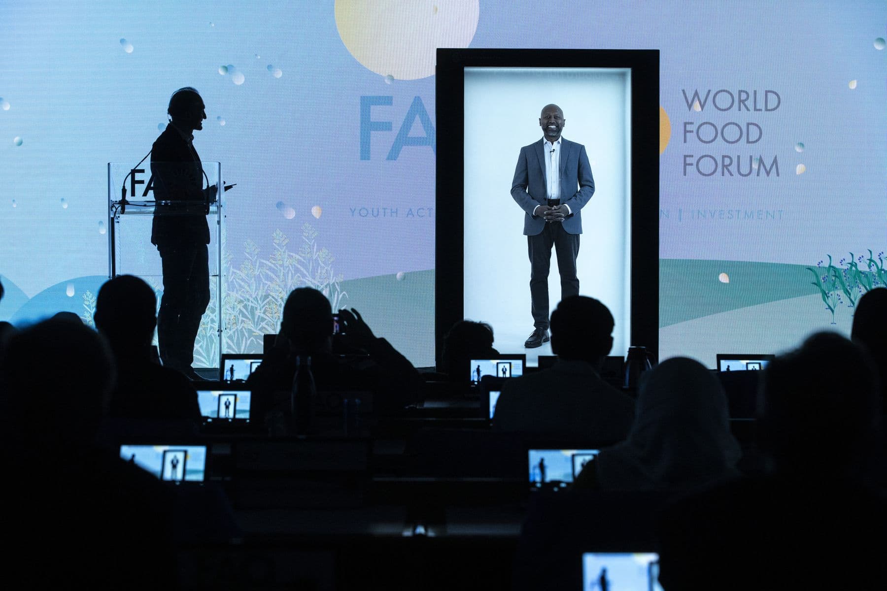 A speaker appearing as a life-size hologram in a Holobox on the FAO World Food Forum stage, with a moderator at the podium.